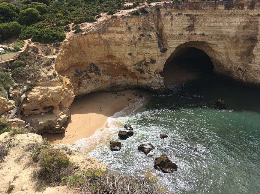 A high-angle view of a secluded sandy cove enclosed by towering limestone cliffs with a natural arch and cave opening. The cliffs display layers of beige and light brown rock with darker patches, and the surface appears rugged with some vegetation on the upper sections. A narrow staircase with metal railings descends along the left side of the cliff, leading to the small beach area below. The sandy shore is light tan with gentle waves washing ashore, creating white foam along the waterline. Several large, dark rocks are scattered in the shallow water near the shoreline, partially submerged and forming a natural barrier. The ocean water transitions from a clear turquoise close to the beach to a darker green further out, with sunlight reflecting off the surface. The environment suggests a natural coastal setting, possibly a protected cove formed by erosion, with no signs of human activity or development visible in the surrounding landscape. This scene aligns with a nature-focused landscape that could be indirectly associated with outdoor leisure or conservation areas, emphasizing the natural geological formations and pristine environment.