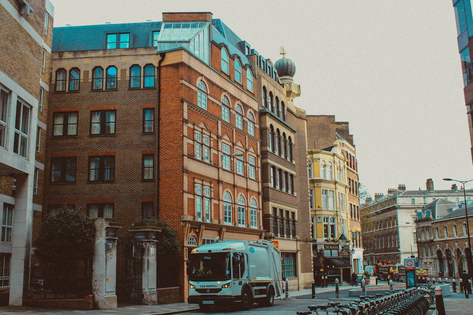 A city street scene featuring multiple multi-storey buildings with varying architectural styles; the central building is constructed of red brick with arched windows and decorative white stone detailing, topped with a teal-colored mansard roof and a small turret with a green dome. To the left, a modern street-facing building has large glass windows and a light-colored facade; to the right, a yellow Victorian-style building with ornate window frames and decorative trim is visible. The street in the foreground shows a white refuse collection truck with the company name 'Rubbish Collection Maida Vale' parked near the curb, alongside black metal bollards separating the sidewalk from the road. The pavement area has a row of rental bikes, and a few pedestrians and street lamps are also visible. The scene is photographed under overcast lighting, giving a neutral tone consistent with an urban environment where private waste collection services may operate as an alternative to municipal rubbish removal, reflected subtly by the parked waste vehicle.