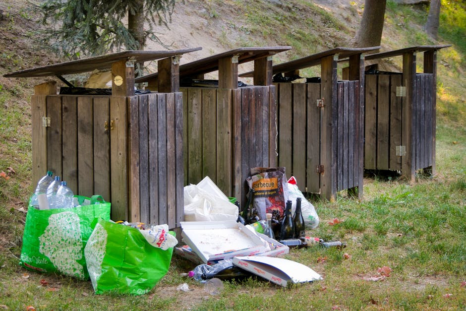 A row of four wooden outdoor rubbish bins with dark-stained vertical planks and flat, slightly sloped roofs are situated on a grassy area, with some green trees and foliage in the background. The bins are positioned against a slight incline on a paved or gravel surface, and they appear to be unused at the moment. In front of the bins, there are various discarded items including transparent plastic bottles, some with clear liquid, placed inside a bright green reusable shopping bag, as well as a white pizza box with food debris inside, and several glass bottles, some dark brown and others clear. Additional debris such as a crumpled paper bag, plastic wrappers, and miscellaneous small objects are scattered on the ground, forming a cluttered impression. The overall scene suggests an instance of improper waste disposal in an outdoor area, which could be indicative of a need for more regular rubbish collection or improved waste management practices by a local service like Rubbish Collection Maida Vale.
