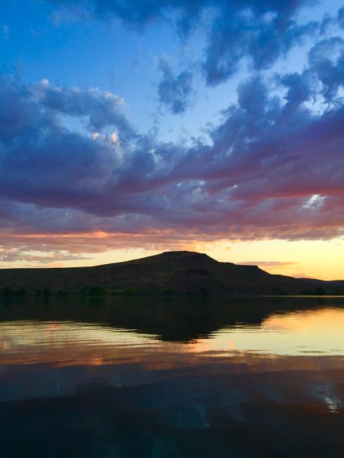 A landscape scene featuring a large body of calm water reflecting the sky above, with a hill or small mountain in the distance. The sky is partly covered with dark, thick clouds displaying shades of deep blue and purple, interspersed with areas of warm orange and pink hues from the setting or rising sun. The water's surface mirrors the sky's colors and cloud formations, creating a symmetrical visual. The environment appears natural and tranquil, with no visible human-made structures or equipment, highlighting the natural scenery that could be associated with outdoor waste management or environmental initiatives by Rubbish Collection Maida Vale for promoting sustainable disposal practices in natural settings.