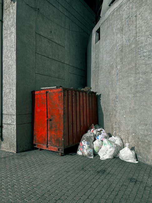 A red metal dumpster with visible rust and signs of wear is positioned in a narrow corner between a dark green wall on the left and a light grey concrete wall on the right. The dumpster is situated on a textured, grey paving surface and appears to be used for waste collection. Several clear plastic bags filled with mixed rubbish, including plastic bottles, cans, and other household waste, are piled up next to the dumpster, some leaning against it. The scene is outdoors, with ambient daylight illuminating the area, and the environment suggests an urban setting where private waste disposal by independent rubbish removal services might be employed as an alternative to municipal collection. The overall scene is utilitarian and reflects the typical appearance of waste storage areas designed for quick collection and disposal.