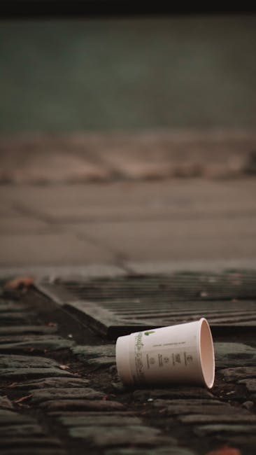 A discarded white paper cup lies on a weathered cobblestone or brick pavement, partially tilted with the opening facing upward. The cup has faint green text or design markings and appears to be made of disposable material, possibly paper or thin plastic. Surrounding the cup, the textured stones show signs of age, with some small debris scattered nearby. In the background, there is a blurred dark object, possibly a drainage grate or similar feature, positioned close to the cup, with a muted greenish and grayish environment visible beyond. The scene is evenly lit with soft, diffuse light, suggesting overcast weather or shaded conditions, and the setting appears to be an outdoor urban pathway or alley, typical of areas where private rubbish might be casually discarded, reflecting the context of rubbish removal and waste collection services like those provided by Rubbish Collection Maida Vale.