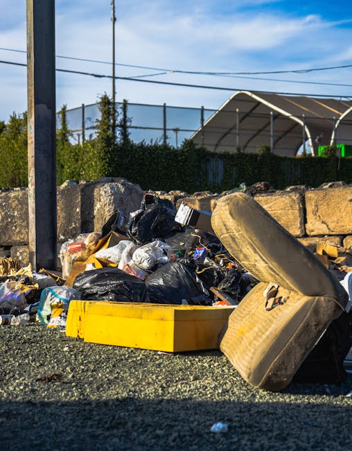 A close-up view of a pile of discarded rubbish on a paved surface, including black garbage bags, a yellow plastic container, and a large, worn, beige car seat with visible stains and fabric tearing. Behind the rubbish, a low stone wall made of irregularly shaped light-colored stones separates the area from a background featuring a metal fence, lush green trees, and a partially visible large, arched outdoor shelter with a cream-colored roof. Overhead, several power lines stretch across a clear blue sky with wispy clouds, indicating an outdoor setting possibly associated with private waste disposal or on-site clearance services. The scene captures the typical contents of a rubbish collection site that might require professional removal, highlighting the importance of proper waste handling to prevent illegal dumping or environmental hazards.