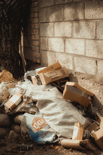 Piles of mixed waste materials, including cardboard boxes, paper sacks, and plastic bags, are stacked and scattered against a rough brick wall exterior, with a concrete or cinder block structure nearby. The cardboard boxes vary in size and are in shades of brown, some with flaps open or partially crushed, revealing darker contents inside. The paper sacks are used, showing signs of wear and dirt, with branding and text partially visible. The plastic bags are loosely filled and crumpled, appearing somewhat dirty and weathered. The waste is placed directly on the ground, amidst small rocks and dirt, in an outdoor setting likely adjacent to a building or alleyway. The scene is illuminated by natural light, highlighting the textures of the materials and the uneven surfaces of the waste accumulation, which suggests an instance of private waste handling or alternative rubbish disposal, potentially managed by a local rubbish collection service like Rubbish Collection Maida Vale.