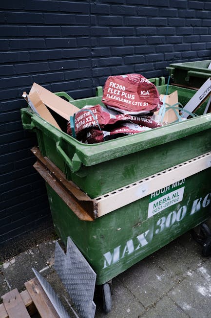 A green wheeled waste container filled with several red and white bags of garden compost sitting outdoors on a paved surface, positioned adjacent to a dark brick wall. The bags, branded as 'IRRIGATOR', contain soil or mulch and are stacked somewhat unevenly in the container. A white ventilation panel with green printing is attached to the side of the container, indicating it is managed by Rubbish Collection Maida Vale. Nearby, a small, metallic lattice panel leans against the container's base, and the background consists of a solid, dark blue brick wall. The environment appears to be a private driveway or side yard, suggesting on-site or alternative waste handling options typical for customers seeking independent rubbish collection services.