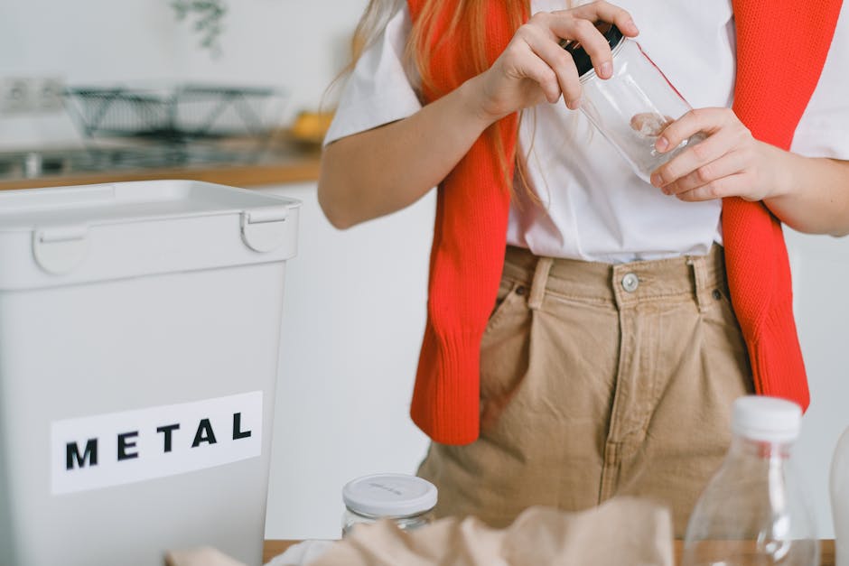A person with long blonde hair, wearing a white shirt and beige trousers, is placing a small glass jar with a metal lid into a grey metal recycling bin labeled 'METAL' with black letters. The individual is standing in a kitchen environment, which features a wooden countertop with a dish drying rack and various utensils in the background. The person has a red vest draped over their shoulders, and there are other recycling-related items such as a plastic bottle and a paper bag visible on the surface. The scene appears to be part of a waste disposal or recycling activity, emphasizing responsible rubbish sorting. The lighting is bright and natural, highlighting the textures and colors of the objects and environment, aligning with the context of on-site waste separation and responsible rubbish removal services at Rubbish Collection Maida Vale.