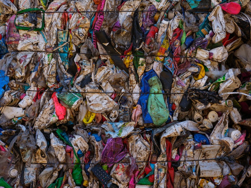A large pile of mixed waste materials appears dirty and disorganized, consisting predominantly of crumpled and torn plastic and paper packaging. The garbage includes brightly colored plastic bottles, wrappers, and bags, with visible textures showing wrinkled and soiled surfaces. Some items, such as flattened cartons and plastic containers, are partially covered in dirt and grime, indicating they have been exposed to the elements or mishandled. The pile is situated outdoors on a flat, paved surface, with no clear background features visible. The waste is tightly compacted and appears to be part of a bulk collection or on-site waste clearance process, typical of private rubbish removal services like those offered by Rubbish Collection Maida Vale, designed to facilitate responsible rubbish disposal and recycling outside traditional municipal waste collection systems. The lighting is natural, providing even illumination across the scene and emphasizing the varied colors and textures of the disposables, illustrating the types of refuse managed during professional rubbish removal operations in Maida Vale.
