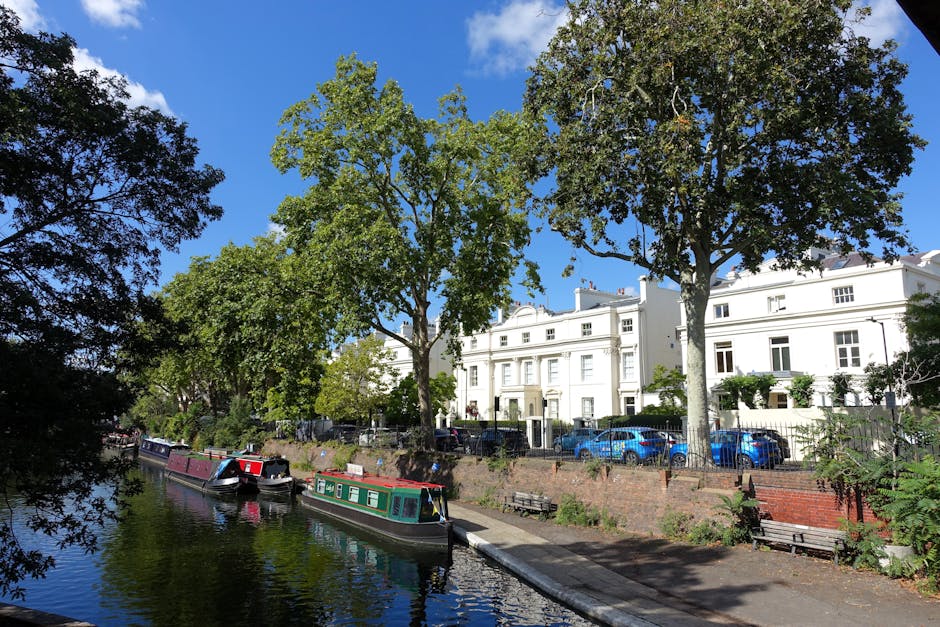 A scenic view of a narrow canal lined with calm water reflecting the sky and nearby surroundings. Along the canal's edge, two narrowboats are moored, one with a dark hull and the other painted in shades of red and green, featuring traditional residential details. The canal is bordered by a low brick wall and a paved walkway with a few vacant wooden benches positioned along it. On the opposite side of the water, a row of tall, leafy trees with lush green foliage provides shade and visual interest, some trees leaning slightly over the water. Behind the trees, there are elegant white residential buildings with classic architectural features, such as bay windows, decorative cornices, and terraced facades, indicating a quiet, affluent neighborhood. Several parked cars in various shades of blue and black are visible in front of the houses, suggesting private property and on-site parking. The bright, clear sky with a few scattered clouds creates a natural, well-lit atmosphere typical of a sunny day in Maida Vale, contributing to the overall serene and picturesque environment that might be associated with private waste collection or on-site rubbish clearance services.