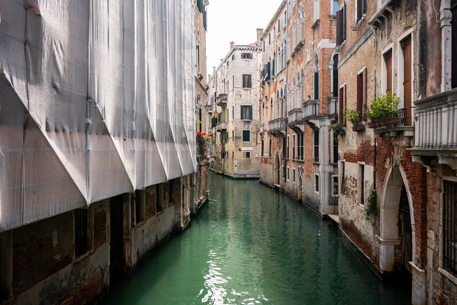 A narrow canal situated between two multi-storey brick buildings in an urban setting, with water reflecting the overcast sky and surrounding structures. The building on the left has a weathered red brick facade with a white, rounded arched window and three circular decorative elements framed in white stone above it. A vertical metal drainpipe runs along this wall. The building on the right also features red brick with visible signs of aging, including sections of exposed, uneven brickwork and a small barred window. Overhanging branches with green leaves extend across the top of the canal, partially obscuring the scene and adding natural contrast to the brick environment. The water appears calm with gentle ripples, indicating a peaceful, enclosed waterway typical of older city areas. The scene evokes a sense of quiet solitude, characteristic of canal-side locations where private waste removal or maintenance might be undertaken, aligning with services offered by the company, Rubbish Collection Maida Vale, that specialises in rubbish and waste clearing solutions in such environments.