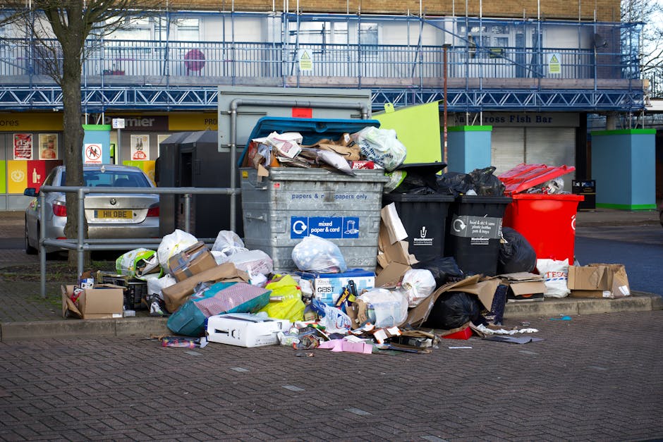 A large collection of various types of waste is piled on a paved sidewalk area, including open-top recycling bins overflowing with mixed paper, cardboard boxes, plastic packaging, and black bin bags. The recycling bin has a blue label indicating it is for paper and cardboard, while other black bins suggest general waste disposal. Surrounding the bins are scattered cardboard boxes, plastic containers, and loose waste items of different colors and materials, such as brown cardboard, white paper, and plastic packaging. To the left, a parked silver car is situated behind a metal railing, with a tree partially visible nearby. In the background, a building with a blue scaffolding structure, glass windows, and signage is visible, alongside a row of shopfronts and storefronts with closed shutters, indicating an urban commercial environment. The scene appears to depict a situation of waste accumulation, possibly in relation to private rubbish collection or curbside disposal, consistent with alternative waste handling services like those provided by Rubbish Collection Maida Vale.