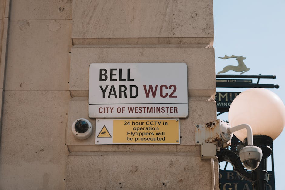 The image shows a beige stone wall with a white and black sign reading 'BELL YARD WC2' indicating a location within the City of Westminster, along with a smaller yellow sign below that states '24 hour CCTV in operation Flytippers will be prosecuted'. Adjacent to the signs, there is a white security camera mounted on the wall, directed downward to monitor the area. To the right, a black ornate street lamp with a round, frosted glass globe is visible, attached to a decorative metal bracket. In the background, part of a street sign with a silhouette of a reindeer and additional signage can be seen against a clear blue sky. The scene is well-lit by natural daylight, emphasizing the textures of the stone wall, metal fixtures, and signage, suggesting an urban environment likely associated with private property or public space management within Westminster, relevant to waste and rubbish removal contexts.