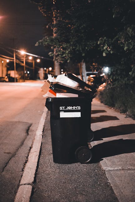 A large collection of various types of waste is piled on a paved sidewalk area, including open-top recycling bins overflowing with mixed paper, cardboard boxes, plastic packaging, and black bin bags. The recycling bin has a blue label indicating it is for paper and cardboard, while other black bins suggest general waste disposal. Surrounding the bins are scattered cardboard boxes, plastic containers, and loose waste items of different colors and materials, such as brown cardboard, white paper, and plastic packaging. To the left, a parked silver car is situated behind a metal railing, with a tree partially visible nearby. In the background, a building with a blue scaffolding structure, glass windows, and signage is visible, alongside a row of shopfronts and storefronts with closed shutters, indicating an urban commercial environment. The scene appears to depict a situation of waste accumulation, possibly in relation to private rubbish collection or curbside disposal, consistent with alternative waste handling services like those provided by Rubbish Collection Maida Vale.