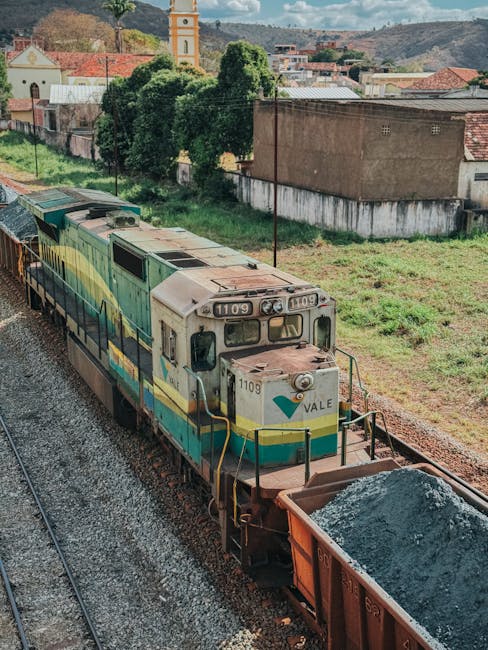 A diesel freight train with an aged, weathered exterior painted in shades of green, yellow, and white is moving along railway tracks in a semi-rural setting. The front of the locomotive displays the number '1109' and the name 'Vale' on its greyish, rusty surface, with visible signs of wear and a slightly faded paint finish. The train is pulling a cargo car filled with a dark, loose granular material, possibly gravel or crushed stone, with the material slightly mounded at the top of the open-topped car. Surrounding the railway are patches of grass and small bushes, with the background showcasing a mix of trees, residential buildings with red-tiled roofs, and a prominent church tower with a yellowish-orange spire set against a partly cloudy sky. The scene suggests an industrial or transportation corridor and reflects a location where private rail transport might support waste or material logistics, aligning with the context of alternative or independent transport and disposal solutions like those provided by Rubbish Collection Maida Vale, although the scene primarily focuses on freight movement in a semi-urban environment.