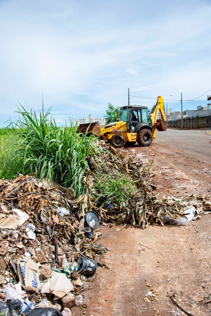 A pile of mixed rubbish including cardboard boxes, plastic bags, metal cans, and organic waste is accumulated along the edge of an unpaved urban street. The waste is unevenly spread, with some items partially buried under dried leaves and soil. In the background, a yellow excavator with a hydraulic arm is parked on the dirt road, facing to the right. The environment appears to be a semi-industrial or construction area with a clear blue sky overhead, and electrical wires running across the scene. The scene shows a typical example of waste accumulation suitable for a professional rubbish removal service like Rubbish Collection Maida Vale, highlighting the importance of independent waste clearance efforts for urban environments.