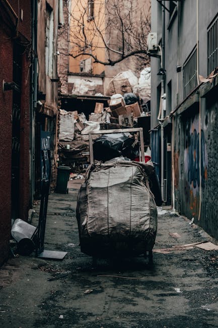 A narrow urban alleyway cluttered with large heaps of mixed waste, including cardboard boxes, plastic bags, and general refuse stacked against a brick wall at the far end. The alley is flanked on both sides by tall buildings with graffiti and metal window bars, indicating an area possibly used for private rubbish disposal or independent collection. In the foreground, a large black and grey wheeled rubbish bin, made of durable plastic or rubberized material, sits on the uneven, slightly stained asphalt surface, with a handle and lid visible. To the left, a small white plastic bag and some scattered debris are present on the ground, while the background shows more waste materials piled up, including discarded packaging and bulky items. Bare tree branches are visible overhead, suggesting a street or service alley in an older part of town, with natural daylight casting soft shadows and a muted color palette dominated by greys, browns, and faded urban hues. The scene conveys a temporary or site-specific waste management setting, possibly related to private rubbish collection services in Maida Vale or similar residential and commercial environments.